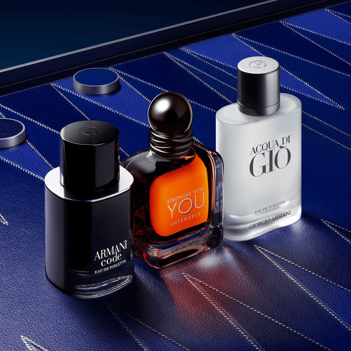Un trio sofisticado de iconos masculinos de Armani: Armani Code, Stronger With You Powerfully y Acqua di Gio. Los frascos sobre un fondo de cuero azul profundo con motivos geometricos couture, encarnando una elegancia masculina absoluta.
