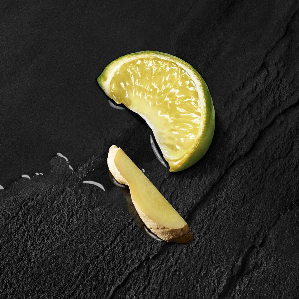 A slice of lime and a piece of ginger resting on a dark, textured surface, with a small amount of liquid visible.