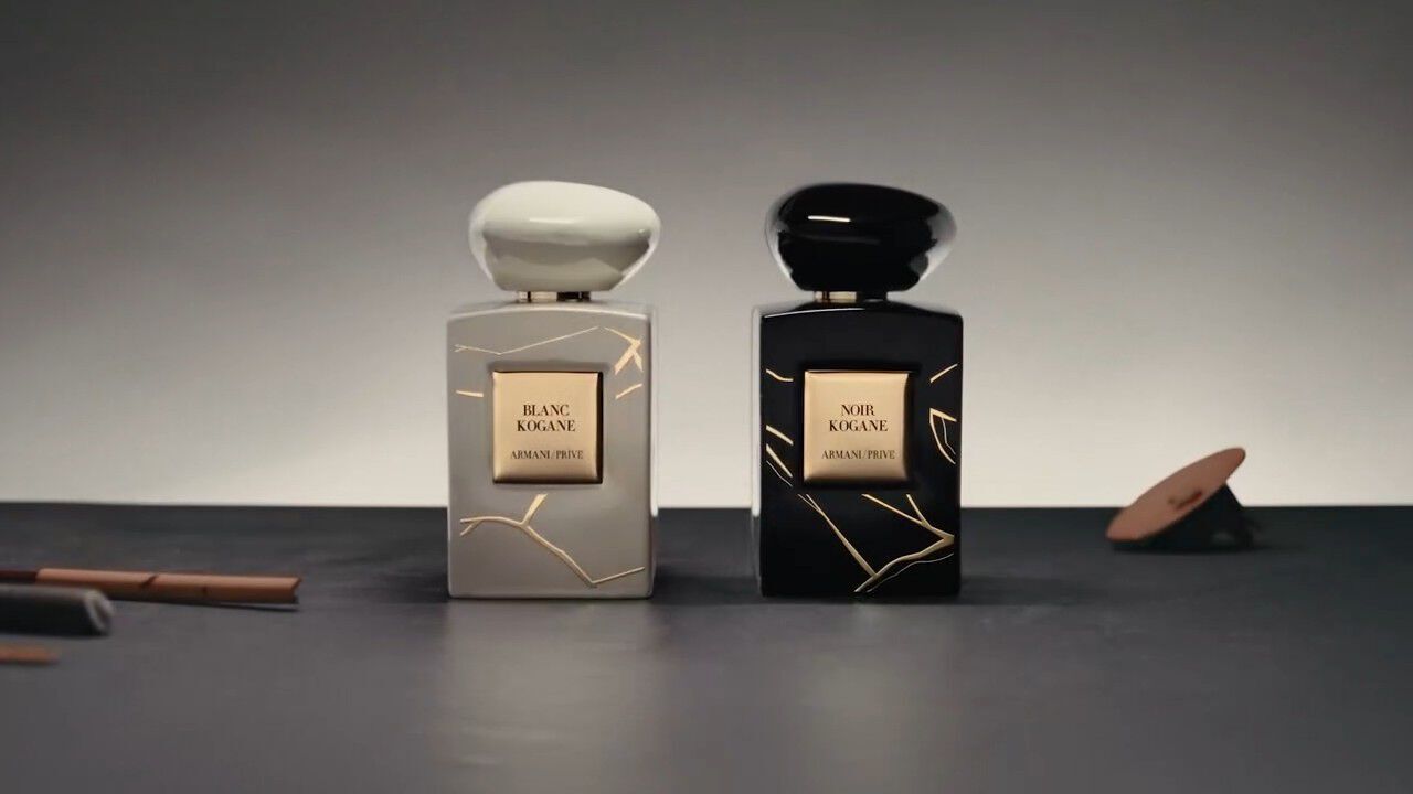 Two elegant perfume bottles, one in white labeled "Blanc Kogane" and the other in black labeled "Noir Kogane," are placed on a dark surface with a minimalist background. Gold accents decorate the bottles.