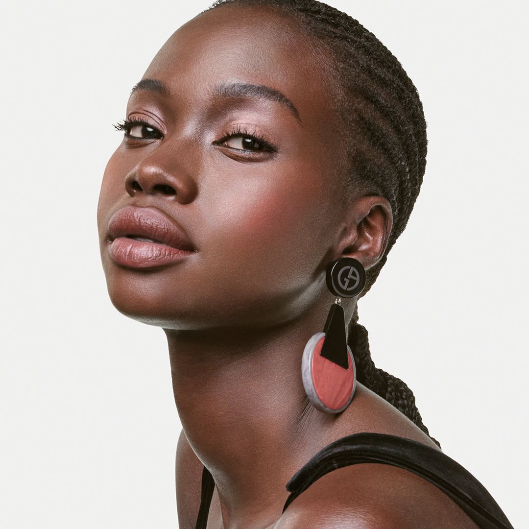 A close-up of a model with braided hair, showcasing bold, artistic earrings and subtle makeup, posed against a light background.