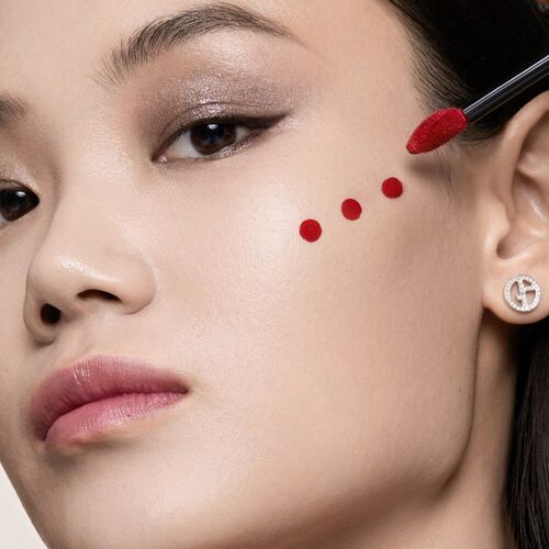 A close-up portrait of a woman with smooth skin and subtle makeup, featuring red dots on her cheek applied with a makeup brush.