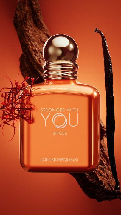 A perfume bottle for EMPORIO ARMANI STRONGER WITH YOU SPICES scent, featuring a warm, amber-toned bottle with a metallic cap. It is artfully arranged with vanilla pods and saffron threads against a vibrant red-orange background, highlighting the aromatic spices.