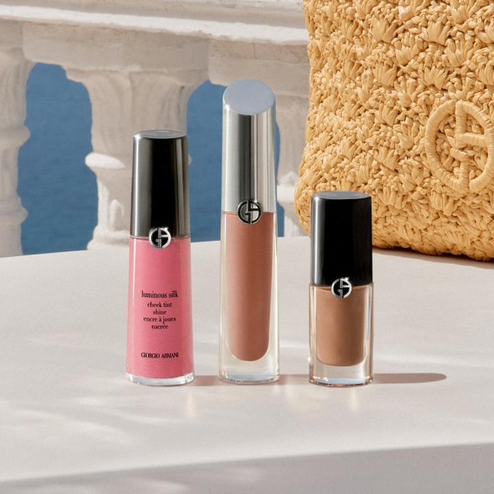 Prodotti di make-up Giorgio Armani esposti su un piedistallo bianco: un blush liquido rosa, un gloss nude e un rossetto liquido marrone, in un’ambientazione d’ispirazione mediterranea con archi in pietra, cielo azzurro e un accessorio dorato dalla texture materica.