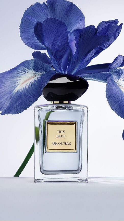 Armani/Prive Iris Bleu: un flacone in vetro trasparente con tappo in pietra nera, davanti a un vibrante iris blu, che cattura un delicato equilibrio tra eleganza floreale e profondita.