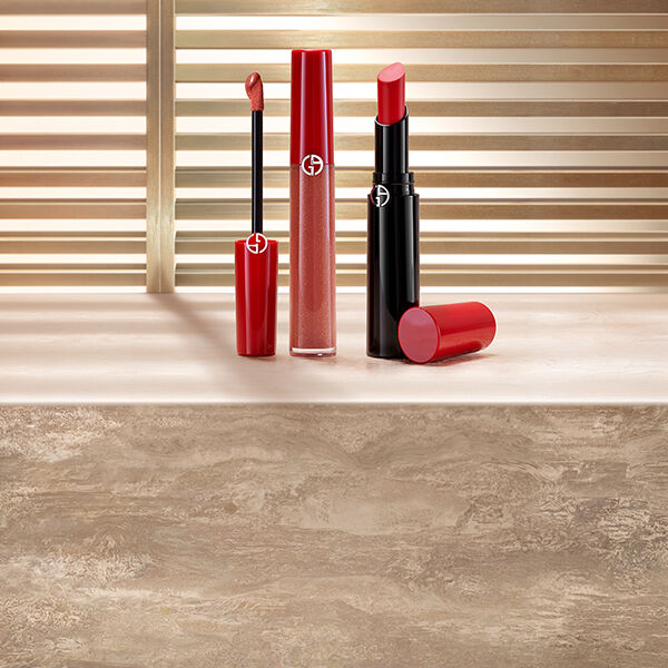 Rossetto liquido rosso e rossetto rosso Giorgio Armani, esposti su una superficie in pietra davanti a veneziane illuminate dal sole.