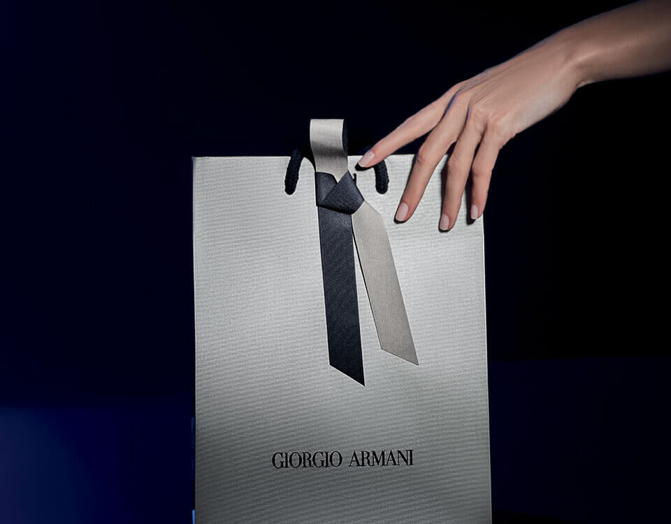 A hand gracefully reaches for a stylish Giorgio Armani gift bag, adorned with a black and grey ribbon on a dark backdrop.