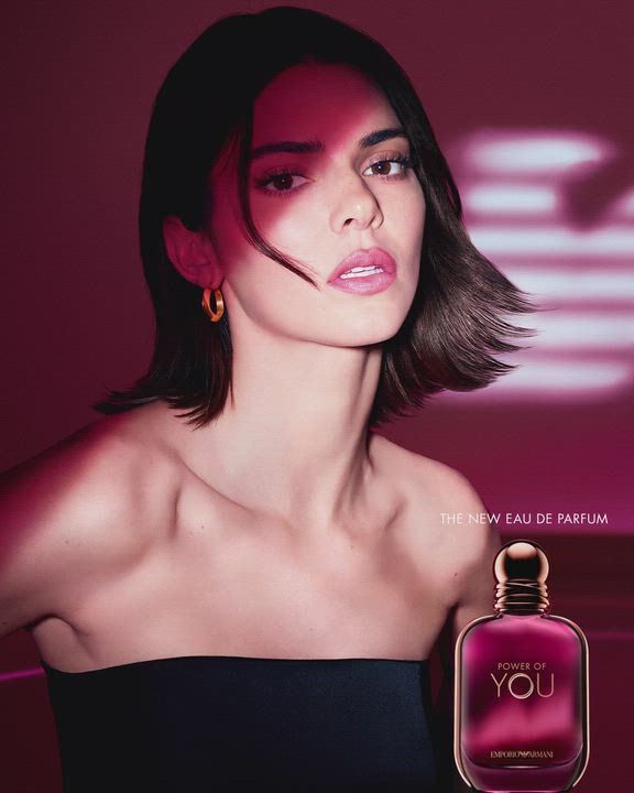 Una pubblicita per l'Eau de Parfum "Power of You" di Emporio Armani. L'immagine ritrae la modella Kendall Jenner a sinistra, che guarda verso l'obiettivo con un'espressione dolce. Ha i capelli corti scuri e indossa un top nero senza spalline e orecchini a cerchio dorati. A destra, e raffigurata una boccetta di profumo color viola-rossastro intenso con la scritta "Power of You" ed "Emporio Armani". Sullo sfondo, il logo dell'aquila di Armani stilizzato e luminoso brilla su una base magenta scuro. Il testo recita "POWER OF YOU" in alto a destra e "THE NEW EAU DE PARFUM" in basso a destra.