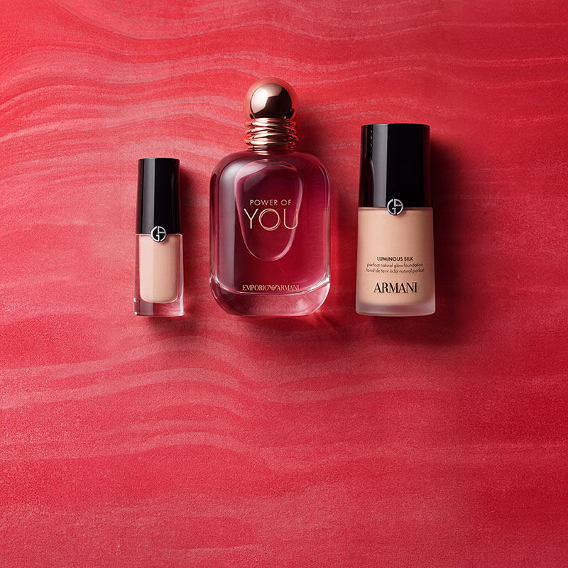 Eine rote Armani Beauty Tasche, umgeben von Duft-, Make-up- und Pflegeprodukten, vor einem strukturierten roten Hintergrund.