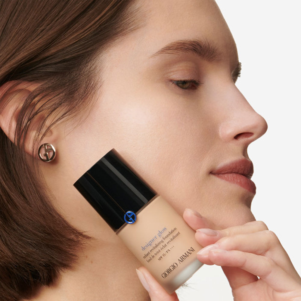 A close-up of a woman holding a bottle of Giorgio Armani foundation, showcasing her clear skin and elegant earrings.