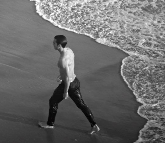 A shirtless man walks along the shoreline, his wet pants glistening in the light as he strides through the gentle waves. The image is in black and white, capturing a serene beach scene.