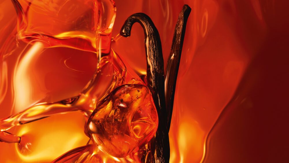 Un primo piano di due baccelli di vaniglia scuri e resina d ambra scintillante su uno sfondo ambrato caldo e luminoso.