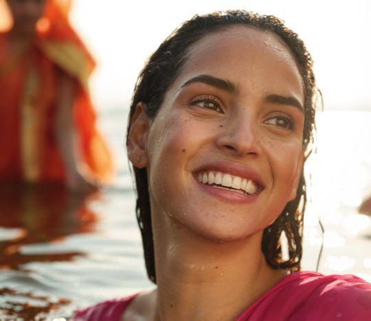 A smiling woman with wet hair is partially submerged in water, wearing a vibrant pink outfit. The sun is setting in the background, creating a warm, golden glow around her.