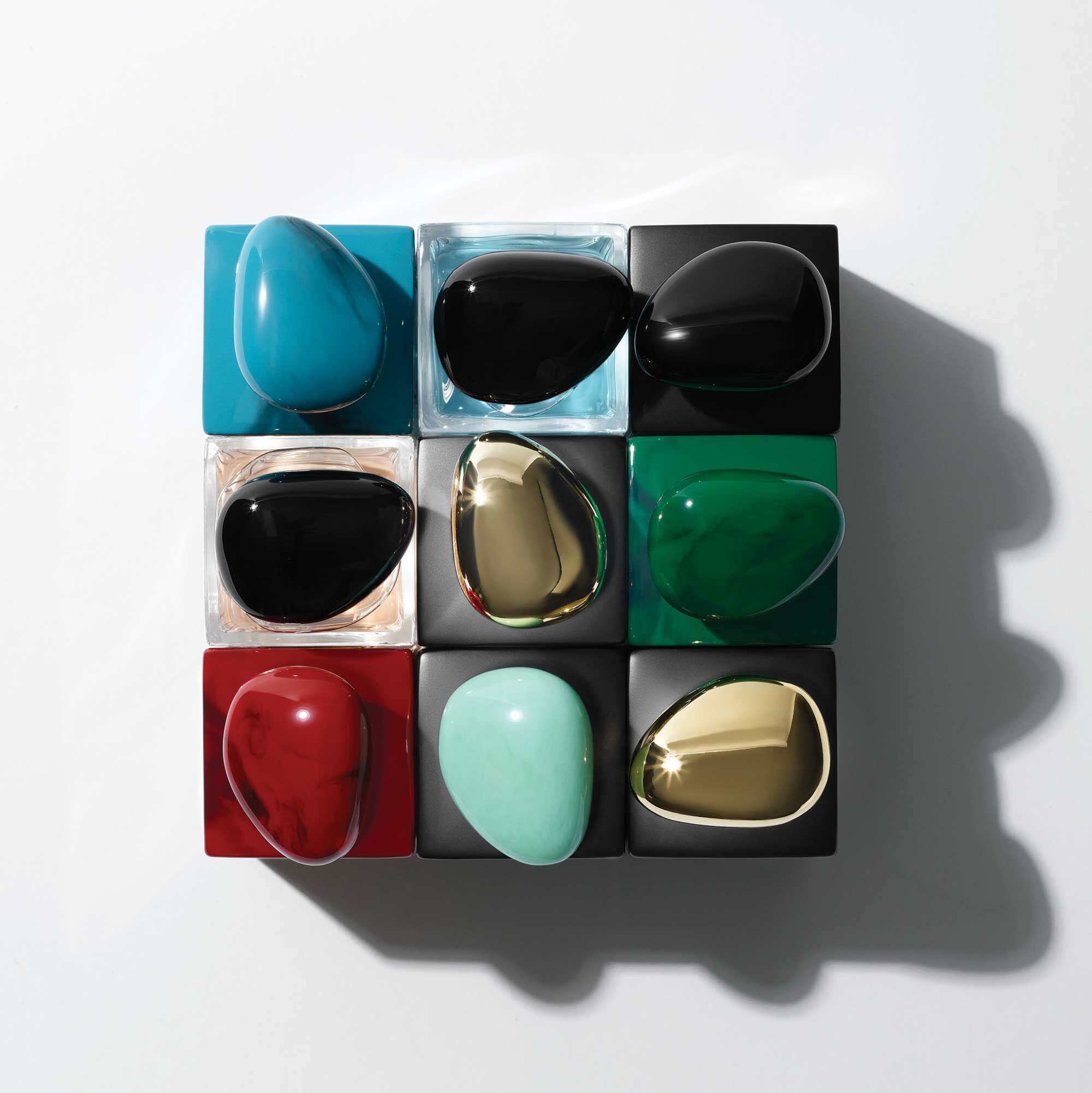A flat lay of nine colorful, smooth, pebble-shaped bottles arranged in a grid. The bottles are in various shades, including turquoise, black, green, red, and gold, against a clean white background.