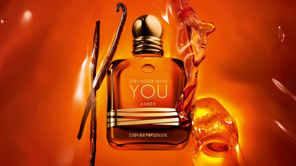 The Emporio Armani Stronger With You Amber fragrance bottle is centrally featured, accompanied by vanilla pods and amber resin, all against a vibrant amber background.