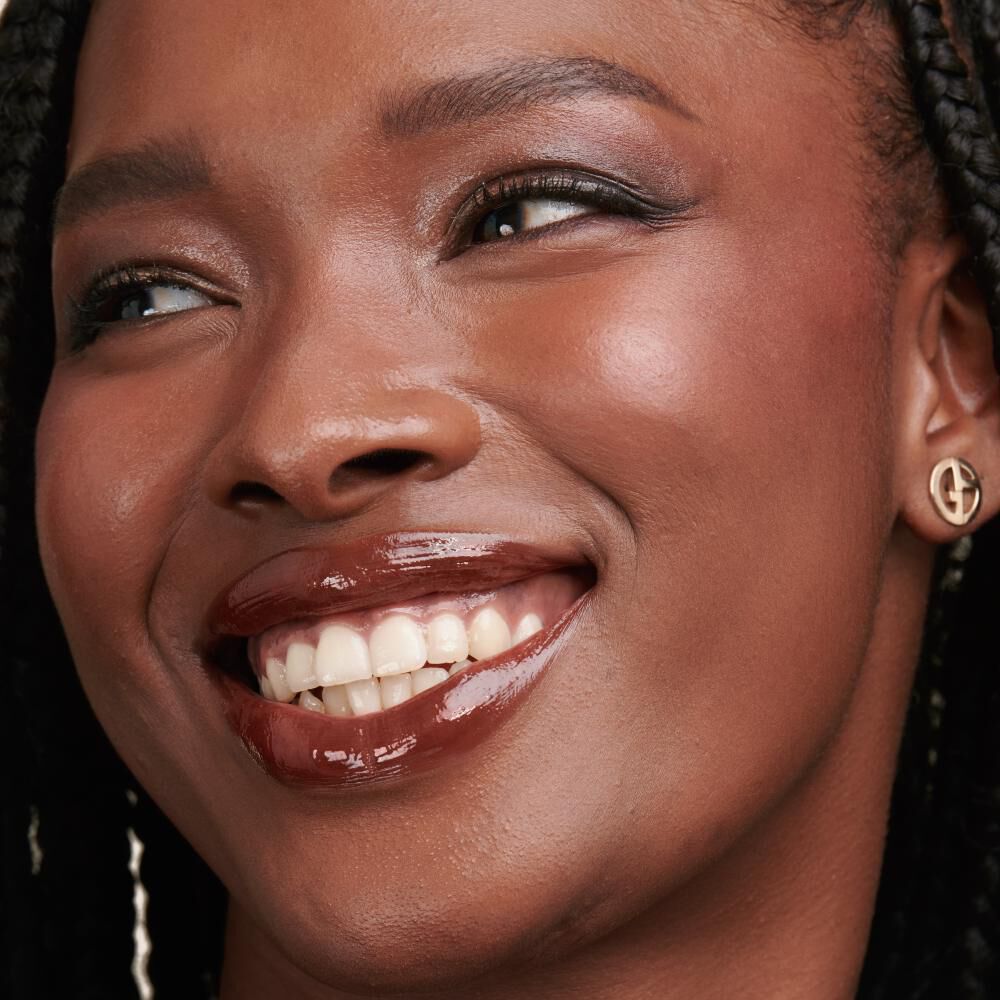 A close-up portrait of a smiling woman with braided hair, showcasing her glowing skin and glossy lips. She wears small earrings and has a radiant expression.