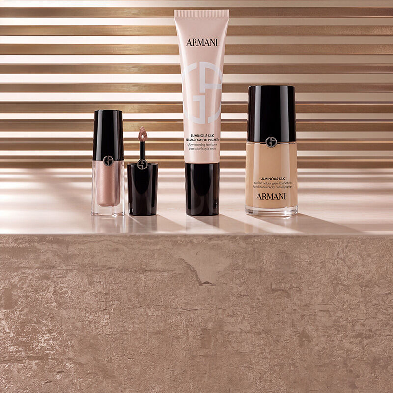  Prodotti di make-up Armani beauty tra cui illuminante liquido, il primer illuminante Luminous Silk e il fondotinta Luminous Silk esposti su una superficie in marmo con uno sfondo caldo a listelli.