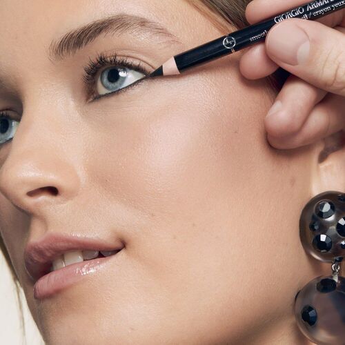 A close-up of a model's face as someone applies eyeliner to her upper eyelid, showcasing her natural beauty with elegant earrings in view.