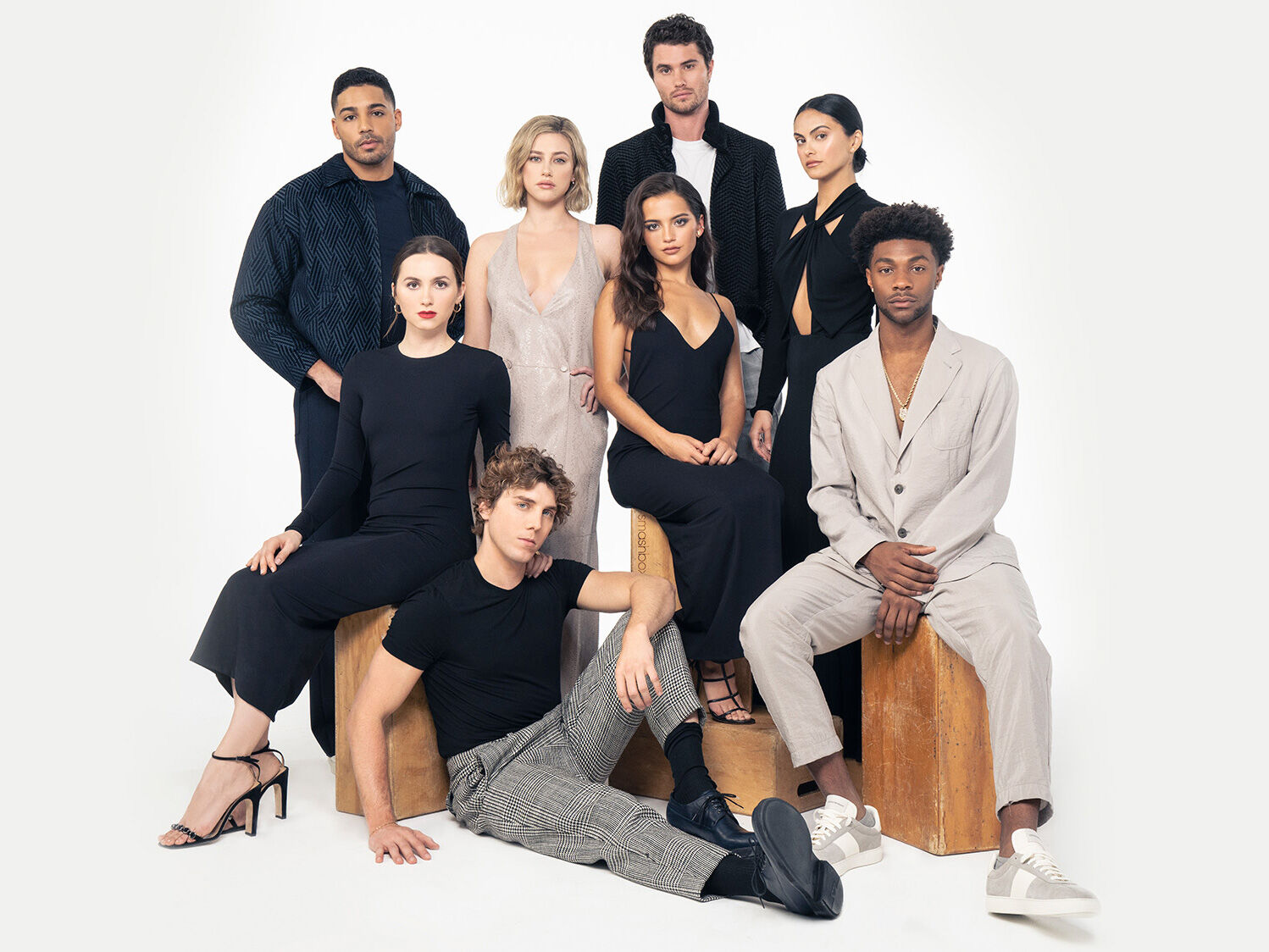 A group of eight individuals poses together in stylish attire, with varying outfits and hairstyles, against a neutral background. They appear confident and are arranged in a mix of seated and standing positions.