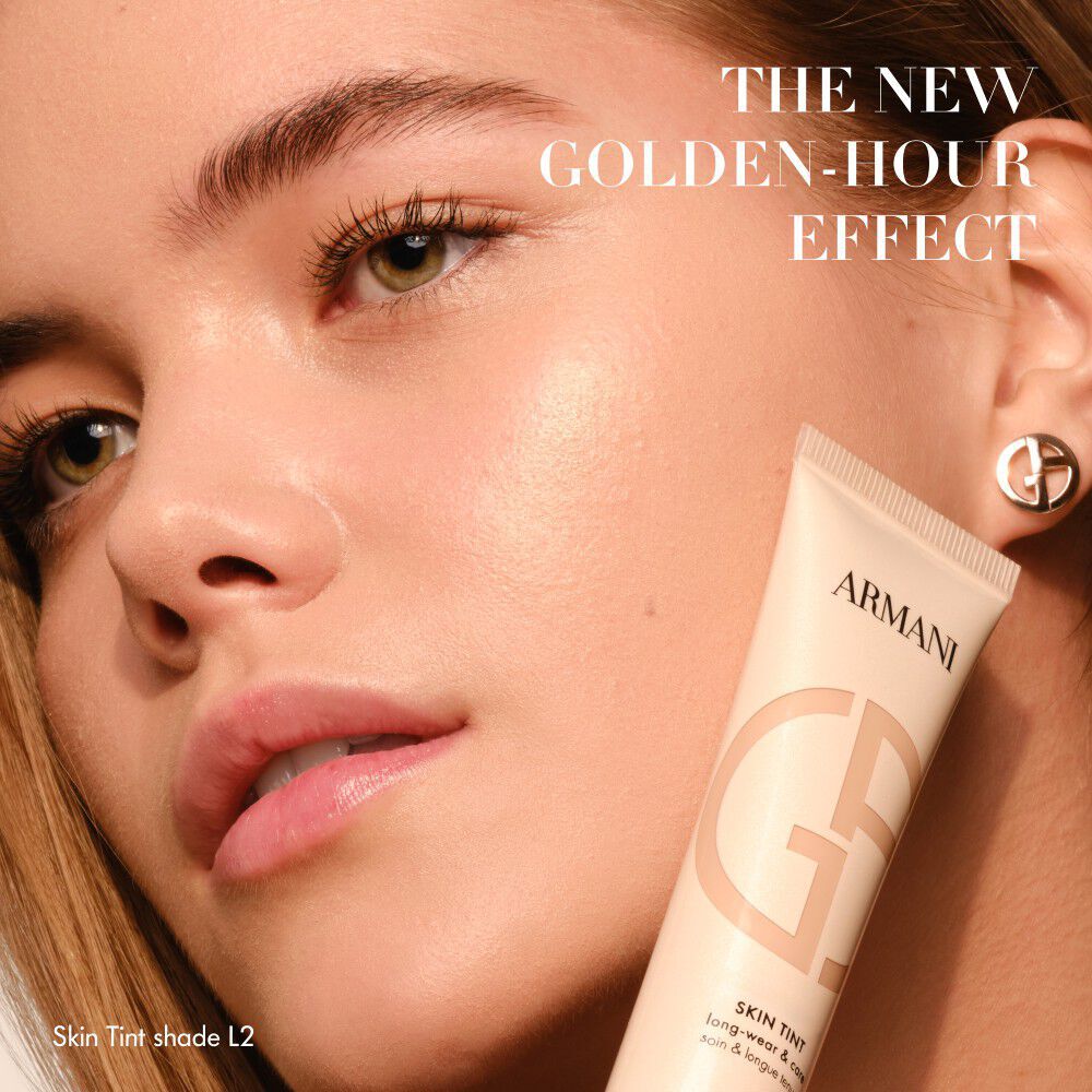 Donna con pelle radiosa che tiene un tubetto di Armani Skin Tint, con la scritta THE NEW GOLDEN-HOUR EFFECT.