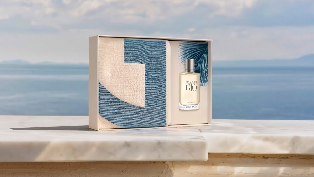 Armani beauty Acqua di Gio Eau de Toilette. The iconic men’s fragrance bottle rests elegantly beside a vintage camera on a blue and white striped canvas, bathed in sunlight. Discover a sophisticated Mediterranean escape.