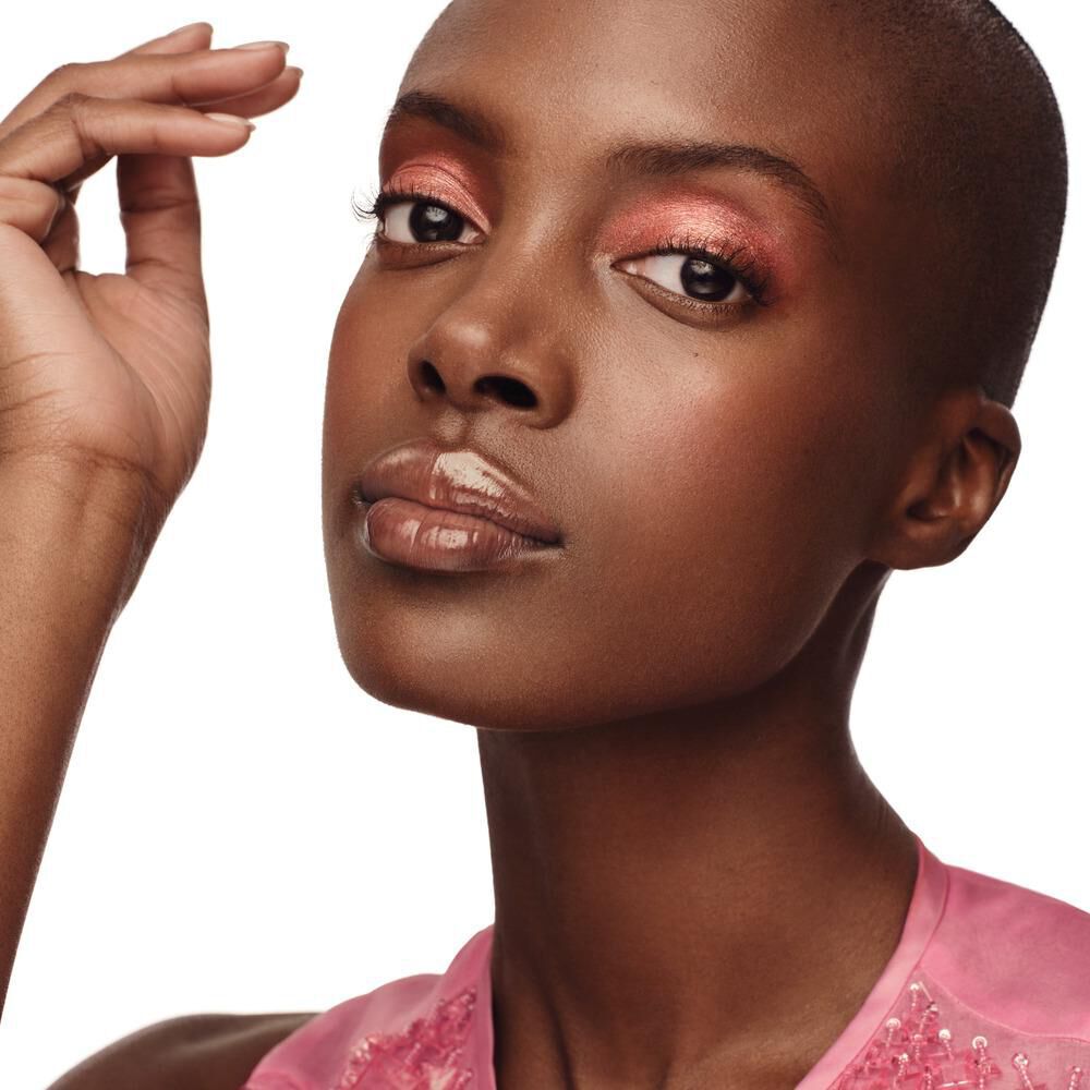 A close-up portrait of a person with a shaved head, showcasing a flawless complexion and vibrant pink eyeshadow, posed with one hand near their face against a white background.