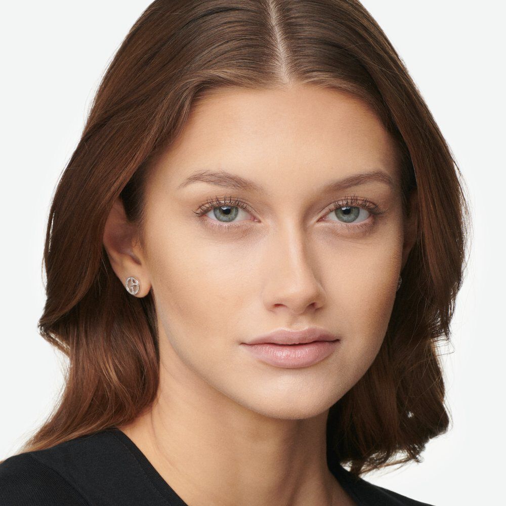 A close-up portrait of a woman with long, brown hair and light makeup, wearing small stud earrings and a black top. She has striking blue eyes and a neutral expression.