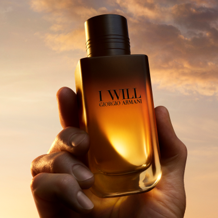 Una mano tiene il flacone di profumo I WILL Giorgio Armani davanti a un cielo dorato al tramonto.