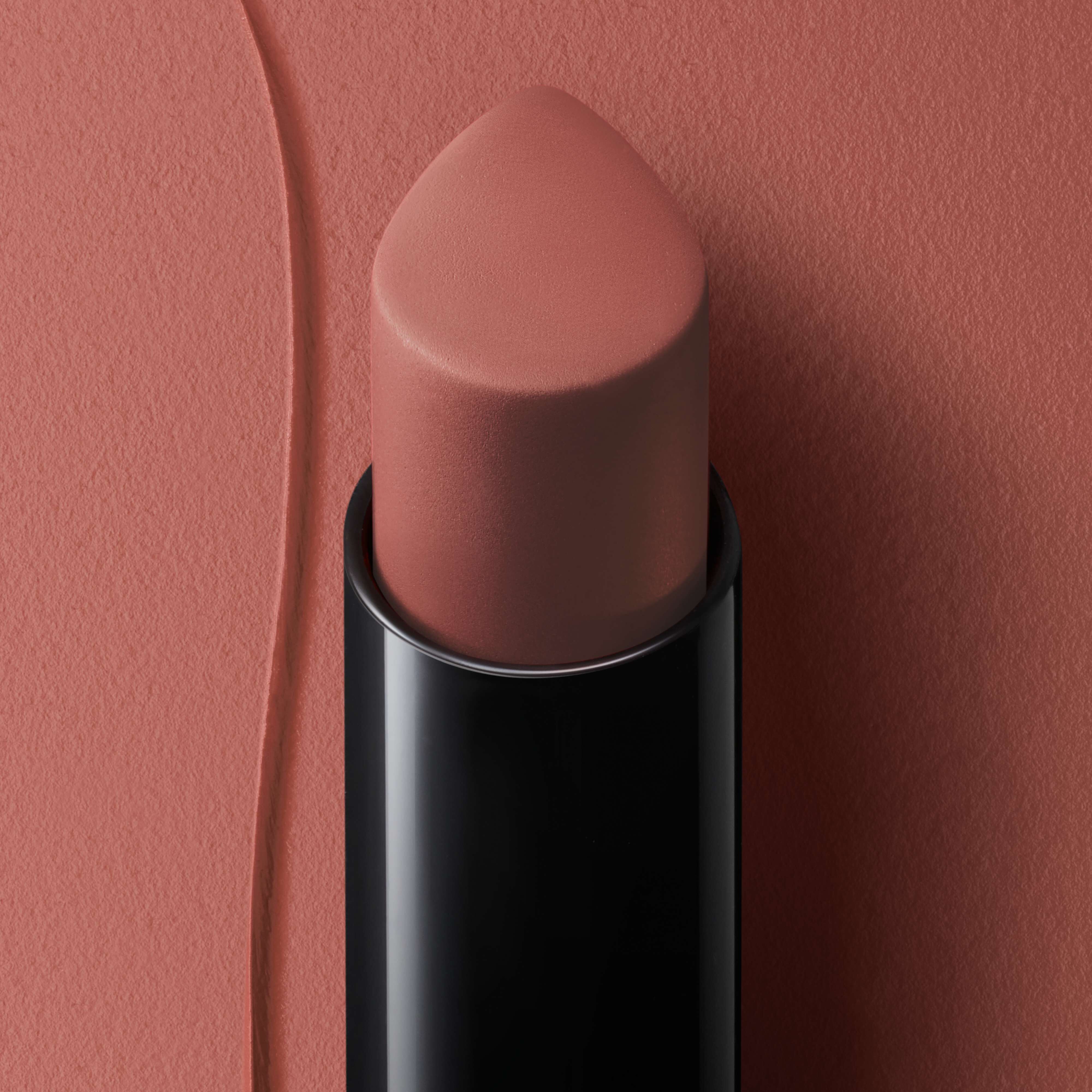 A close-up of a lipstick bullet in a sleek black casing, showcasing a soft, muted nude color against a textured coral background.