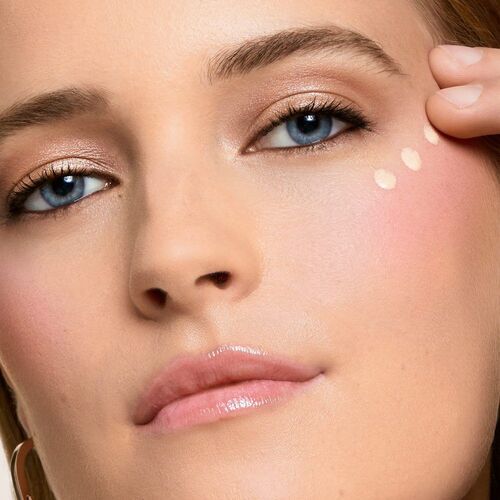 A close-up image of a woman with blue eyes and a natural makeup look, applying a small amount of foundation to her cheek.