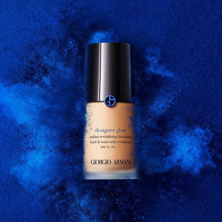A bottle of Giorgio Armani Designer Glow revitalizing foundation, set against a blue powder background, emphasizing the product's vibrant color and luxurious design.