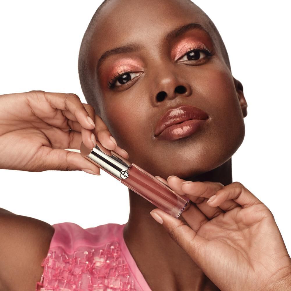 A woman with a bald head and a radiant makeup look holds a lip gloss tube near her face, showcasing a soft, glossy lip color. She wears a pink embellished top and has a confident expression.