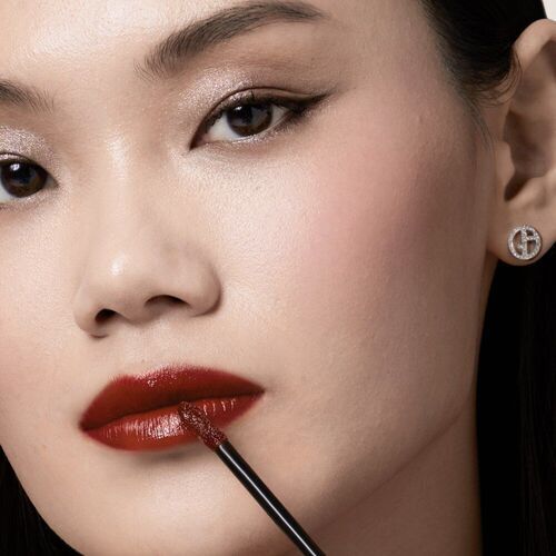 A close-up of a woman applying dark red lip color with a brush, highlighting her defined features and subtle eye makeup. She is wearing diamond stud earrings.
