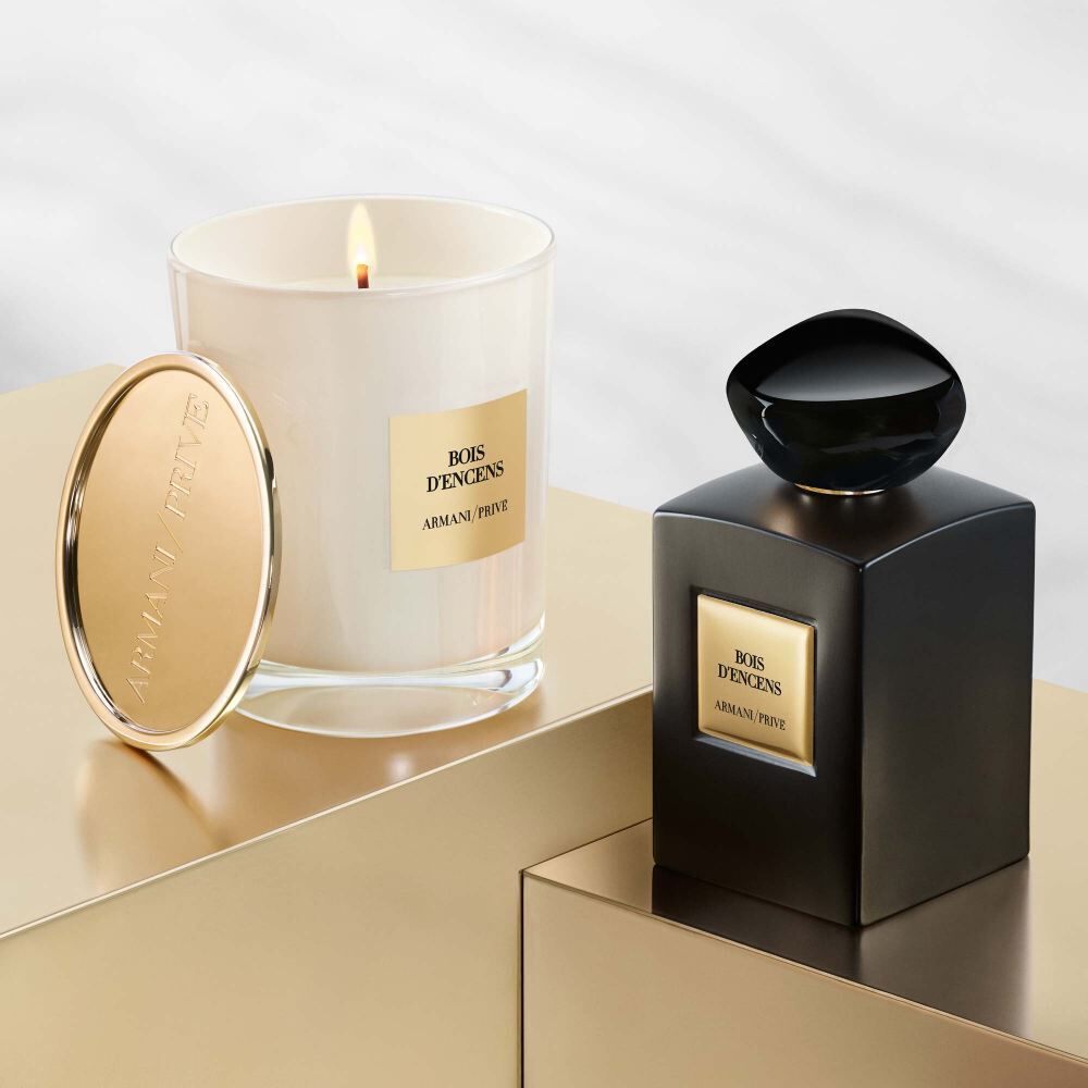 A sleek black perfume bottle and a white candle, both featuring elegant gold labels with the text "BOIS D'ENCENS" by Armani Prive, displayed on a reflective surface.