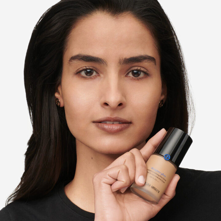 A woman with long dark hair holds a bottle of foundation, looking directly at the camera with a neutral expression. She is wearing a simple black shirt.