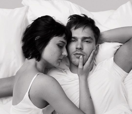 A black-and-white portrait of a woman with short hair and a man lying together on a bed, with the woman resting her head on the man's shoulder and gently touching his face.