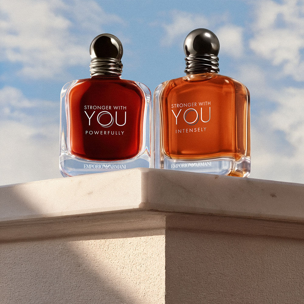 Le fragranze Stronger With You Powerfully e Stronger With You Intensely di Emporio Armani, presentate su un piedistallo in pietra sotto un cielo azzurro.​