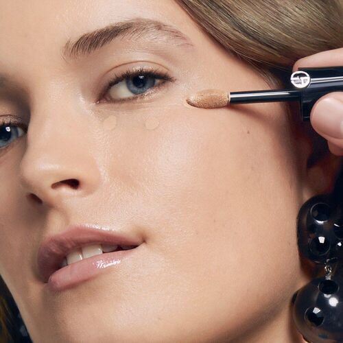A close-up of a woman applying makeup under her eye, with a makeup applicator in hand. She has a natural look, featuring soft skin and minimal makeup, and is wearing large, glossy earrings.