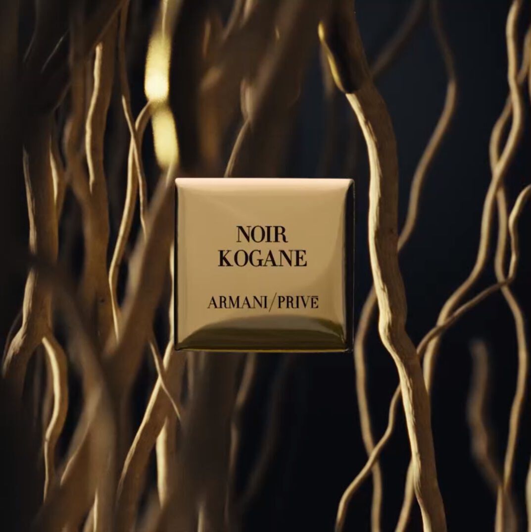 A gold square fragrance bottle labeled "NOIR KOGANE" by Armani Prive, set against a backdrop of intertwining branches or roots.