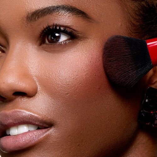 A close-up of a woman applying blush to her cheek with a makeup brush, showcasing her clear skin and bright smile.