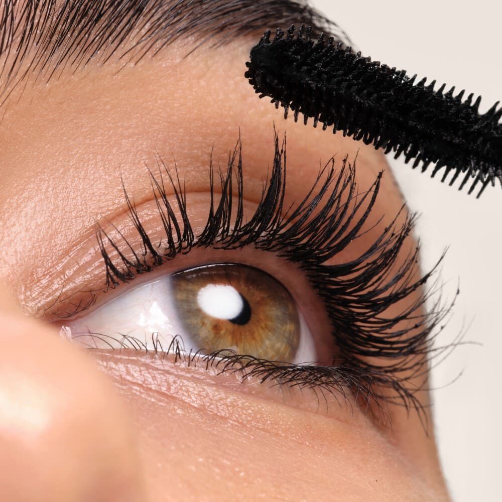 Immagine ravvicinata di un occhio con ciglia nere folte, mentre si applica il mascara. L'iride e marrone chiaro.