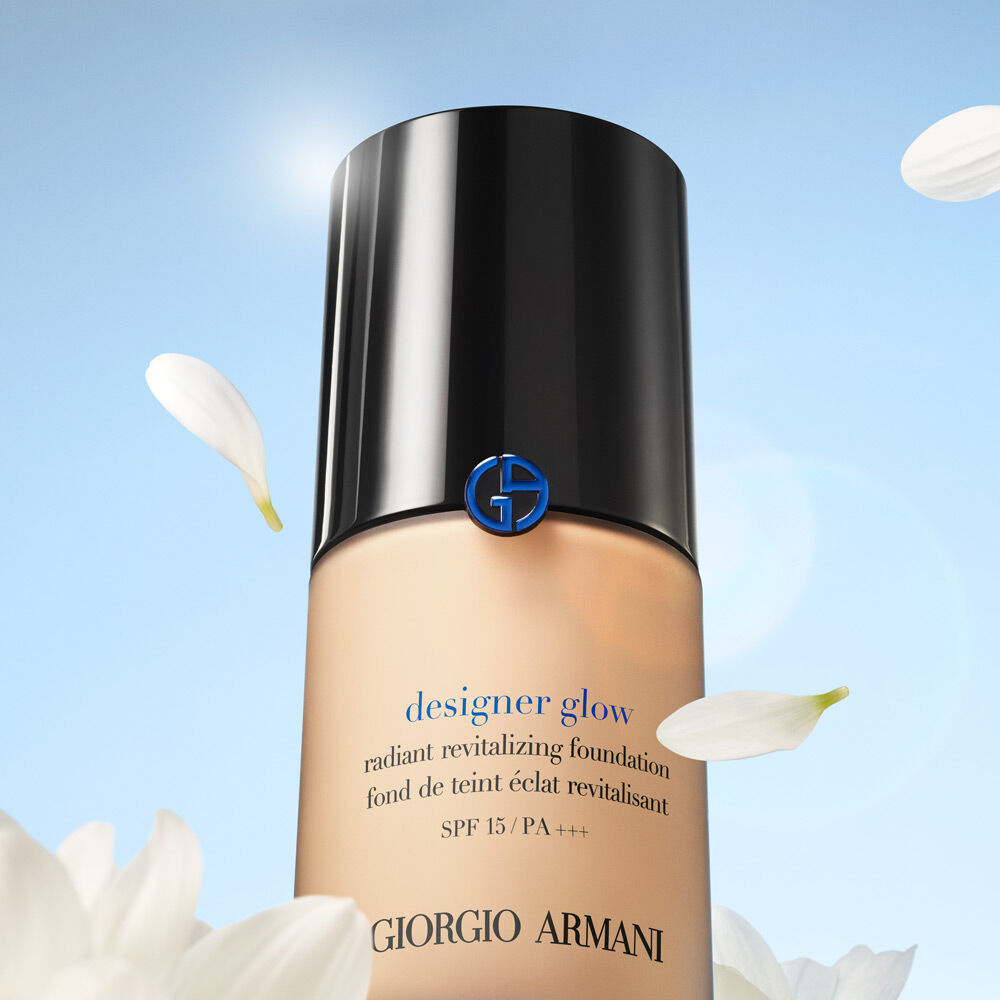A bottle of Giorgio Armani Designer Glow Radiant Revitalizing Foundation, showcasing a sleek design with a black cap and blue logo, surrounded by white flower petals.