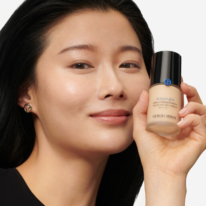 A smiling woman with long dark hair holds a bottle of Giorgio Armani foundation, showcasing her glowing skin.