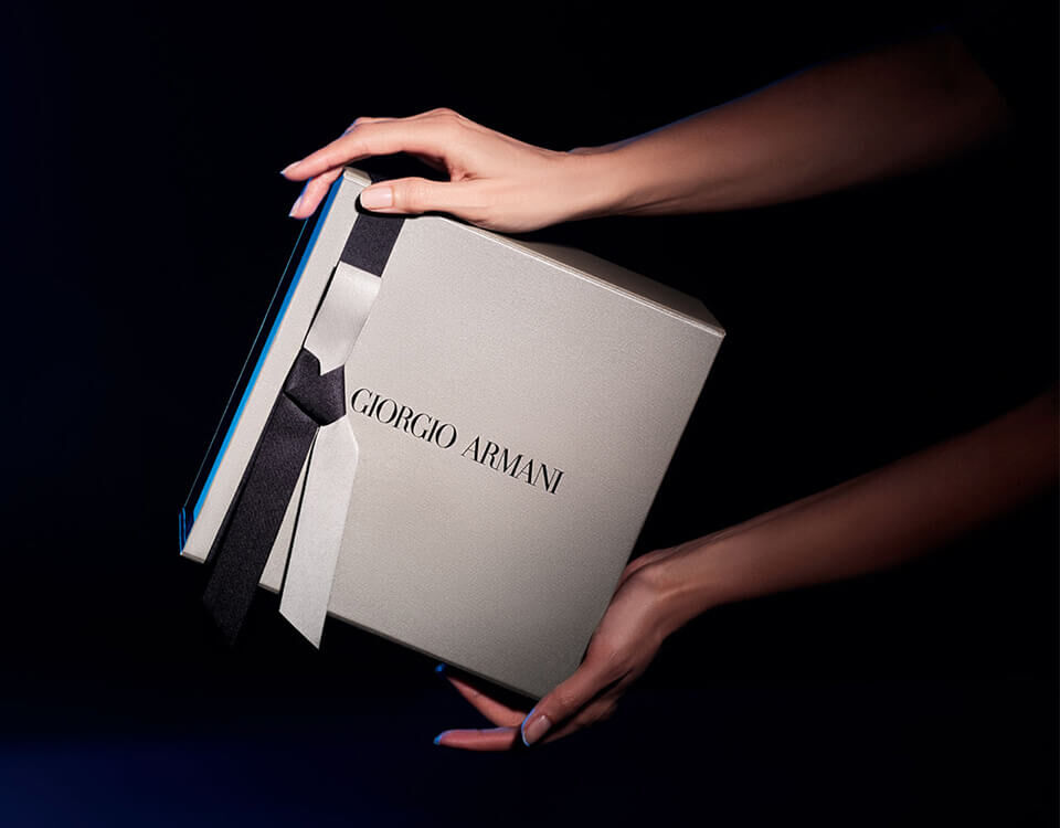 A pair of hands holding a luxurious Giorgio Armani gift box, elegantly tied with a black ribbon, against a dark background.