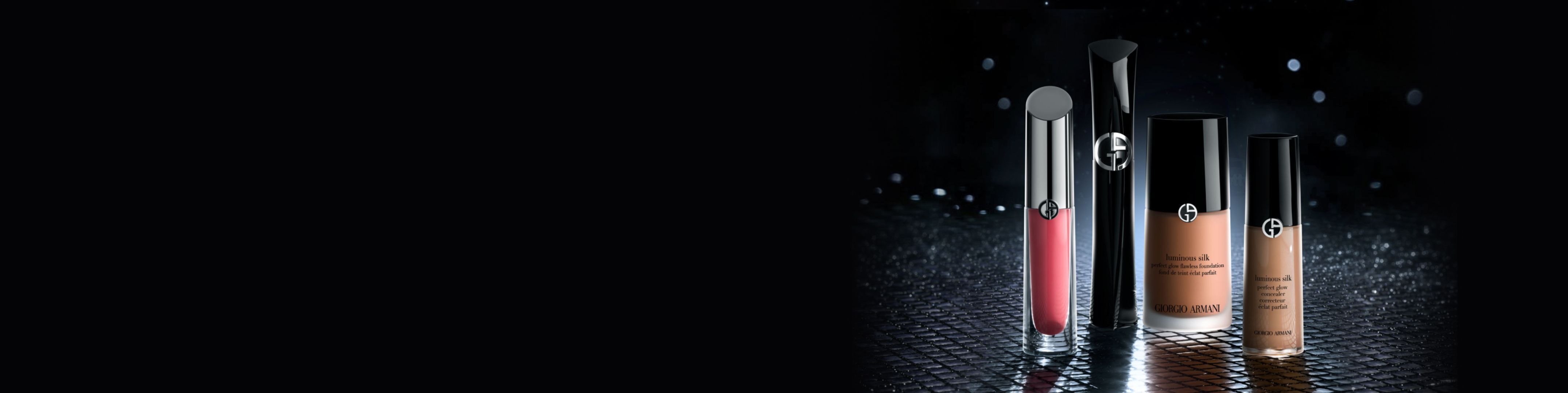 Make-up Armani Beauty su sfondo scuro: lacca labbra rossa, mascara nera, fondotinta Luminous Silk e correttore su superficie riflettente.