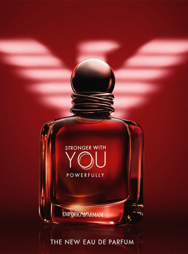 Una pubblicita d'impatto per l'Eau de Parfum "Stronger With You Powerfully" di Emporio Armani. L'immagine mostra una boccetta di vetro squadrata e robusta, riempita con un liquido rosso ambrato intenso che brilla dall'interno. La boccetta ha un tappo scuro e arrotondato con un dettaglio di fili intrecciati intorno al collo. Le scritte "STRONGER WITH YOU POWERFULLY" ed "EMPORIO ARMANI" sono stampate in argento e bianco. Sullo sfondo, un grande logo dell'aquila di Armani stilizzato e illuminato in bianco su una base rosso intenso. In basso, il testo recita "THE NEW EAU DE PARFUM".
