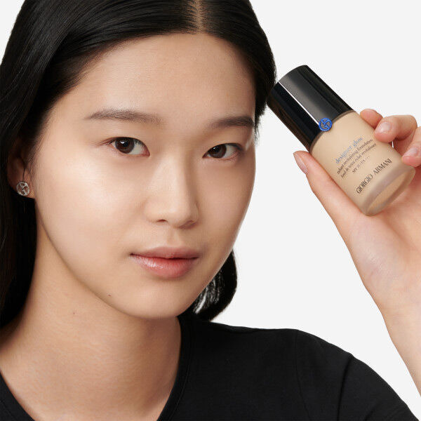 A young person with smooth, natural skin holds a bottle of foundation in one hand, looking directly at the camera with a neutral expression. They have long black hair and are wearing a simple black shirt.