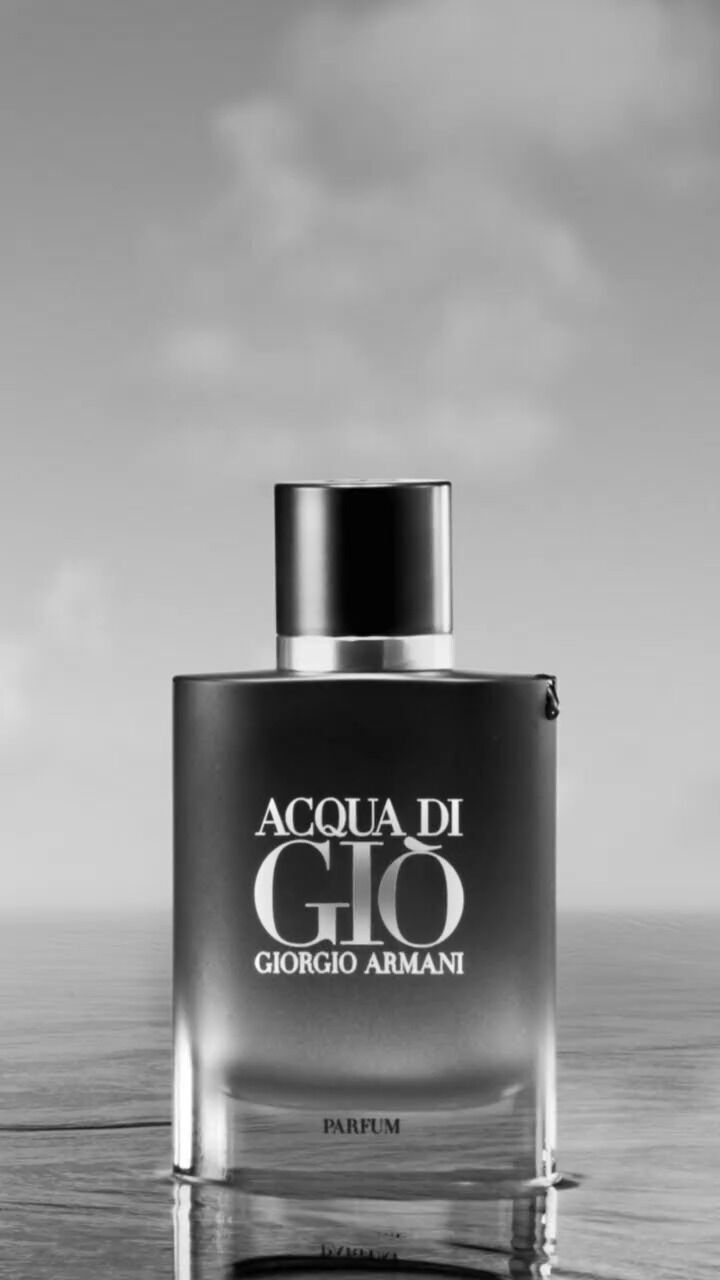 A sleek, clear glass bottle of Acqua di Gio perfume by Giorgio Armani, set against a soft, cloudy background. The bottle features a black cap and the brand name in bold white lettering.