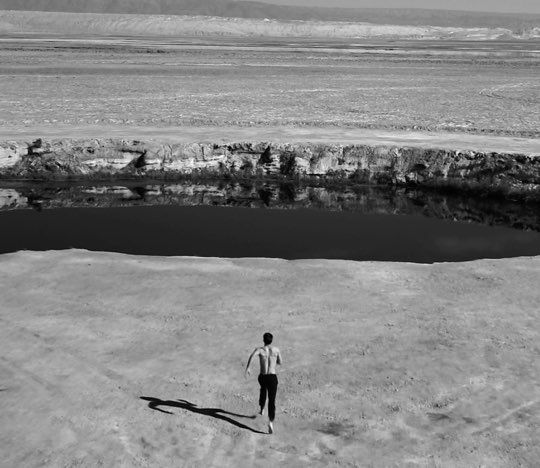 A lone figure walks along the edge of a dry, barren landscape, with a small body of water reflecting the sky. The scene is depicted in black and white, emphasizing the starkness of the environment.