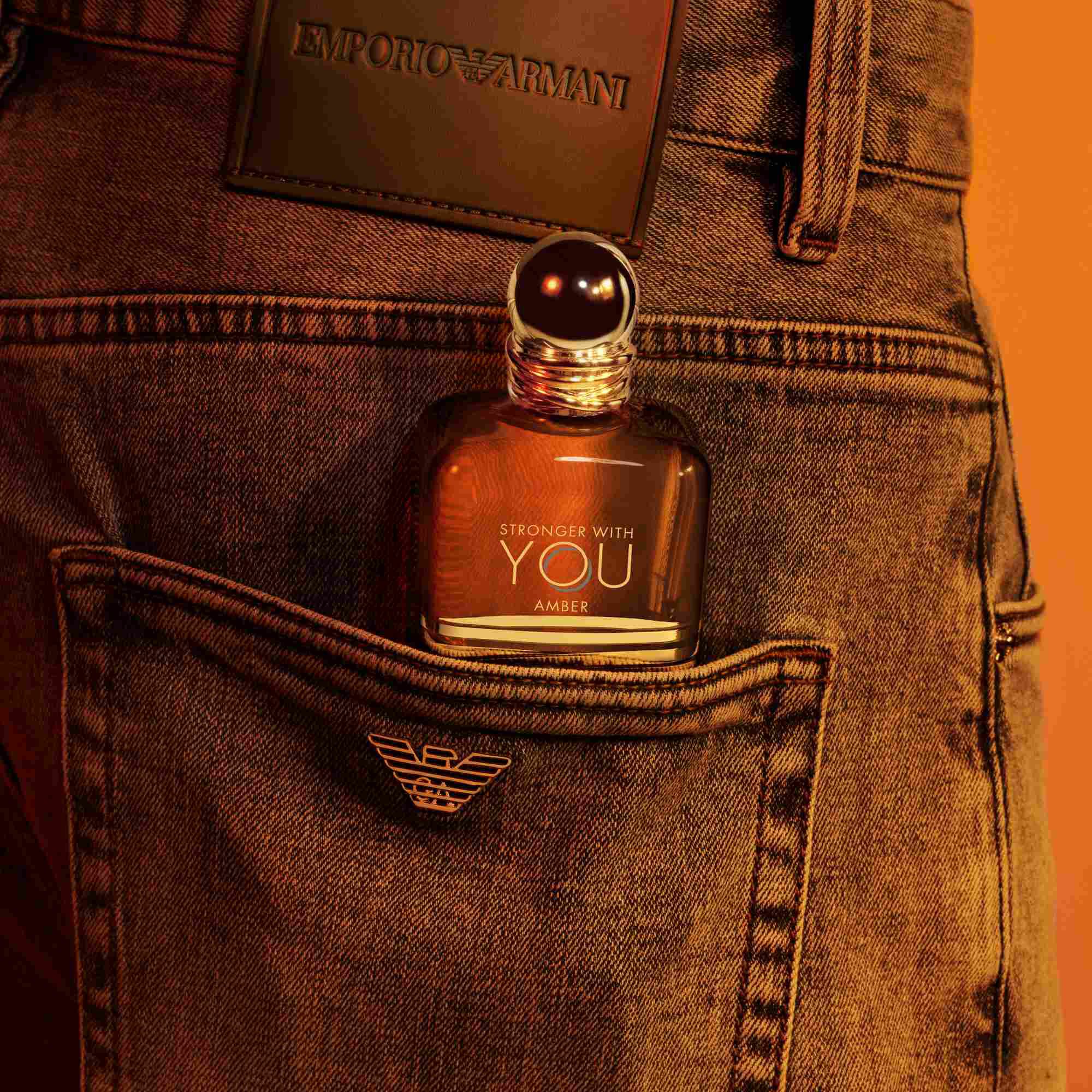 Un flacone di profumo Emporio Armani Stronger With You Amber che spunta dalla tasca posteriore di un paio di jeans, con una toppa in pelle scura visibile sopra